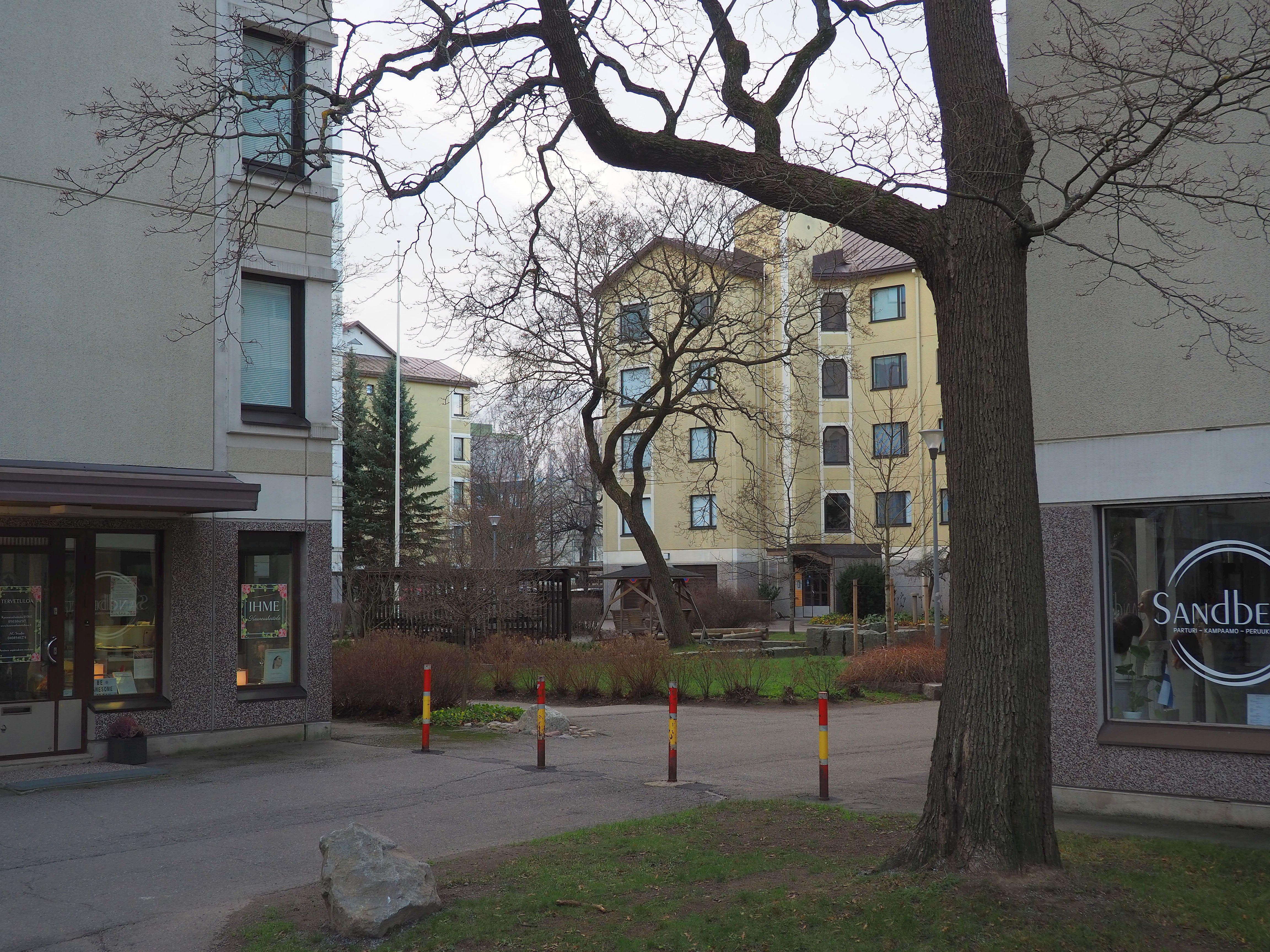 Näkymä korttelin sisälle. Kerrostalojen kivijalkaliikkeitä ja lehdettömiä puita.