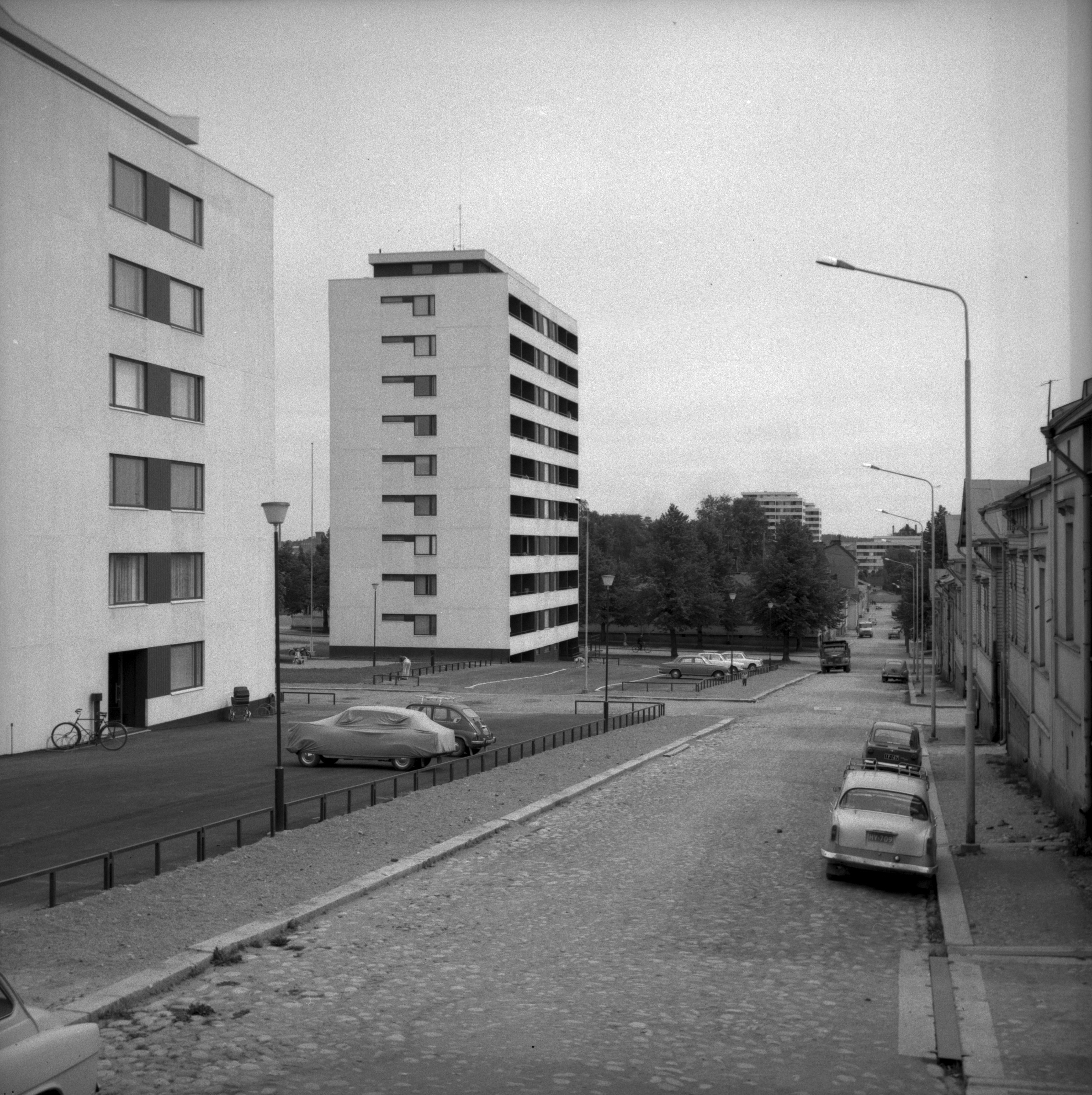 Toisen kerroksen tasalta otettu valokuva katunäkymästä. Kaksi 10-kerroksista kerrostaloa vasemmalla puolella ja puutaloja oikealla, autoja pysäköitynä kadulla.