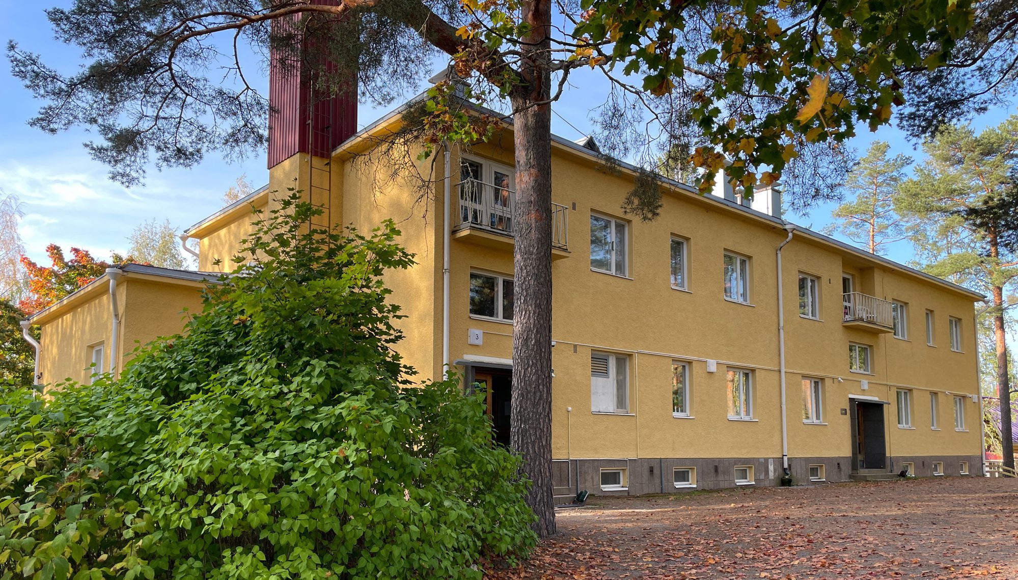 Syksyllä otettu kuva keltaiseksi rapatusta kaksikerroksisesta rakennuksesta. Etualan asvalttipäällysteisessä ylämäessä pudonneita lehtiä.