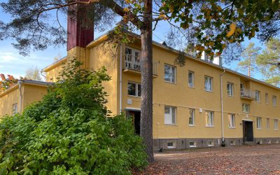 Syksyllä otettu kuva keltaiseksi rapatusta kaksikerroksisesta rakennuksesta. Etualan asvalttipäällysteisessä ylämäessä pudonneita lehtiä.