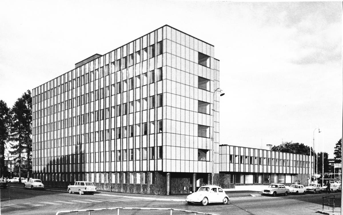 Mustavalkokuvassa kuusikerroksinen virastorakennus, jonka oikealla puolella on kaksikerroksinen siipirakennus. Rakennuksen edessä kadulla on autoja.