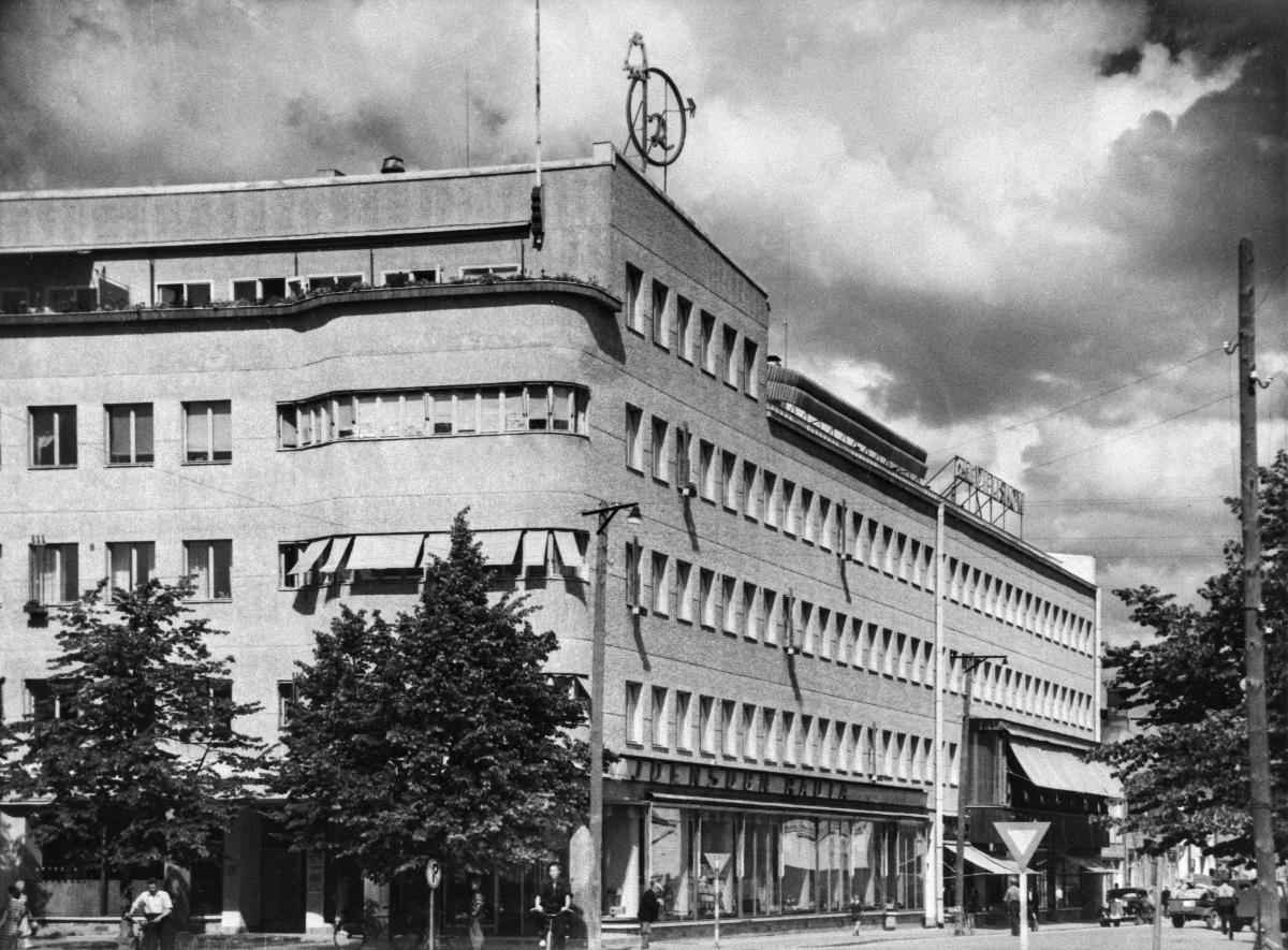Mustavalkokuvassa viisikerroksinen rakennus, jonka alimmassa kerroksessa on liiketiloja.