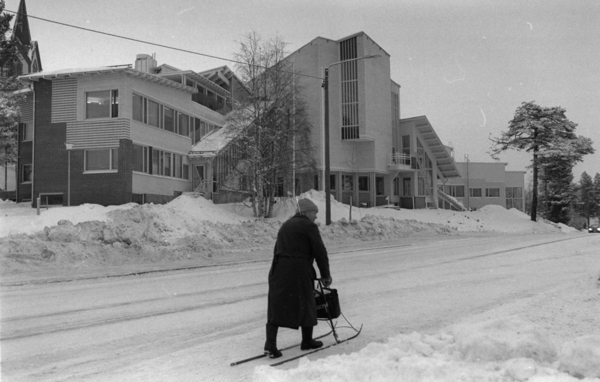 Mustavalkokuvassa talvivaatteisiin pukeutunut henkilö liikkuu potkukelkalla virastorakennuksen edessä.