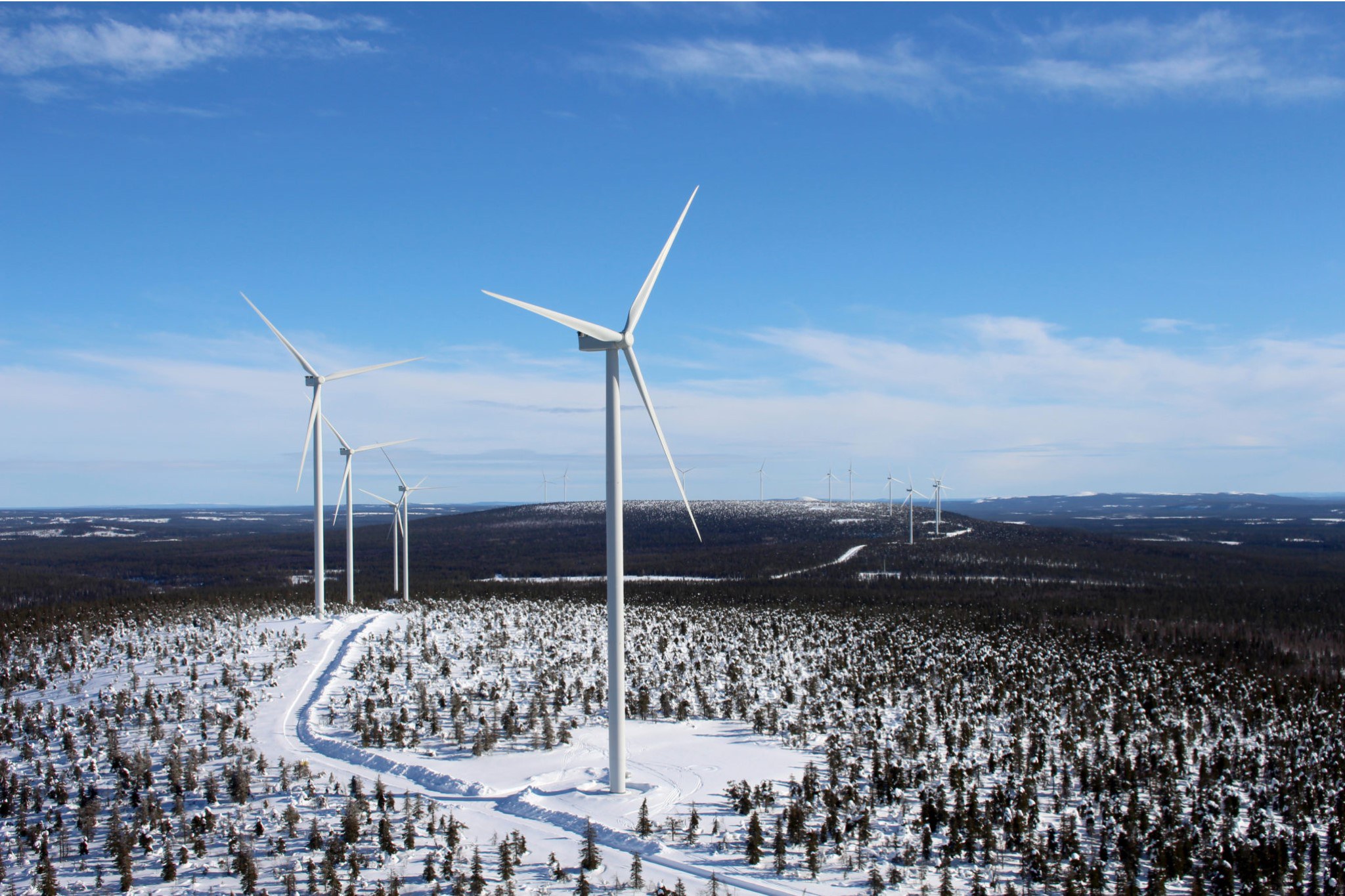 Talvinen ilmakuva kumpuilevasta metsämaisemasta, jossa on useita tuulivoimaloita.