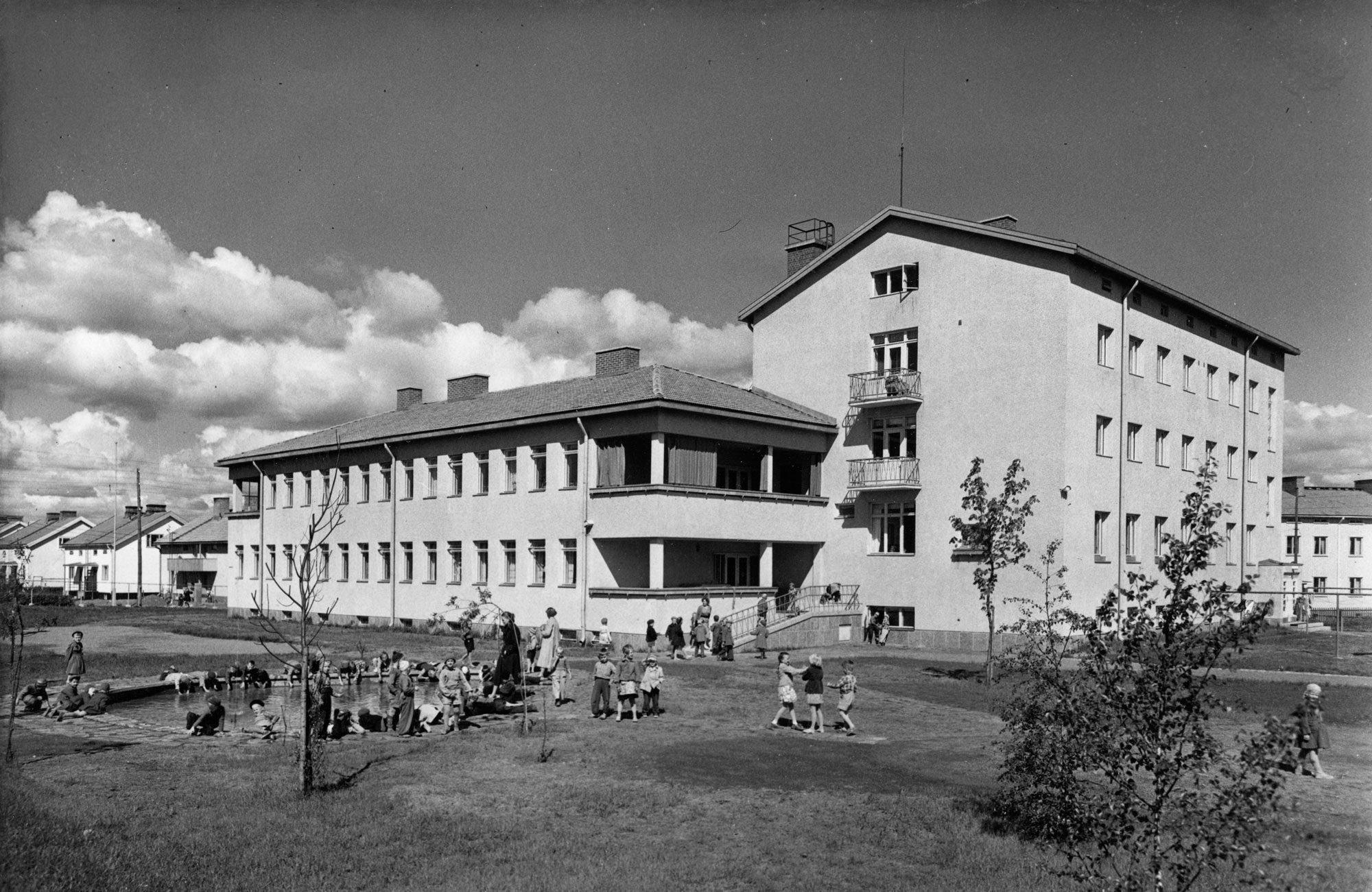 Mustavalkokuvassa on suuri, kolmikerroksinen rapattu rakennus, jossa on harjakatto ja useita rivejä suorakaiteen muotoisia ikkunoita. Rakennuksen edessä on nurmialue, jossa näkyy lapsia leikkimässä ja aikuisia heidän seurassaan. Taustalla on muita asuinrakennuksia ja pilvinen taivas.