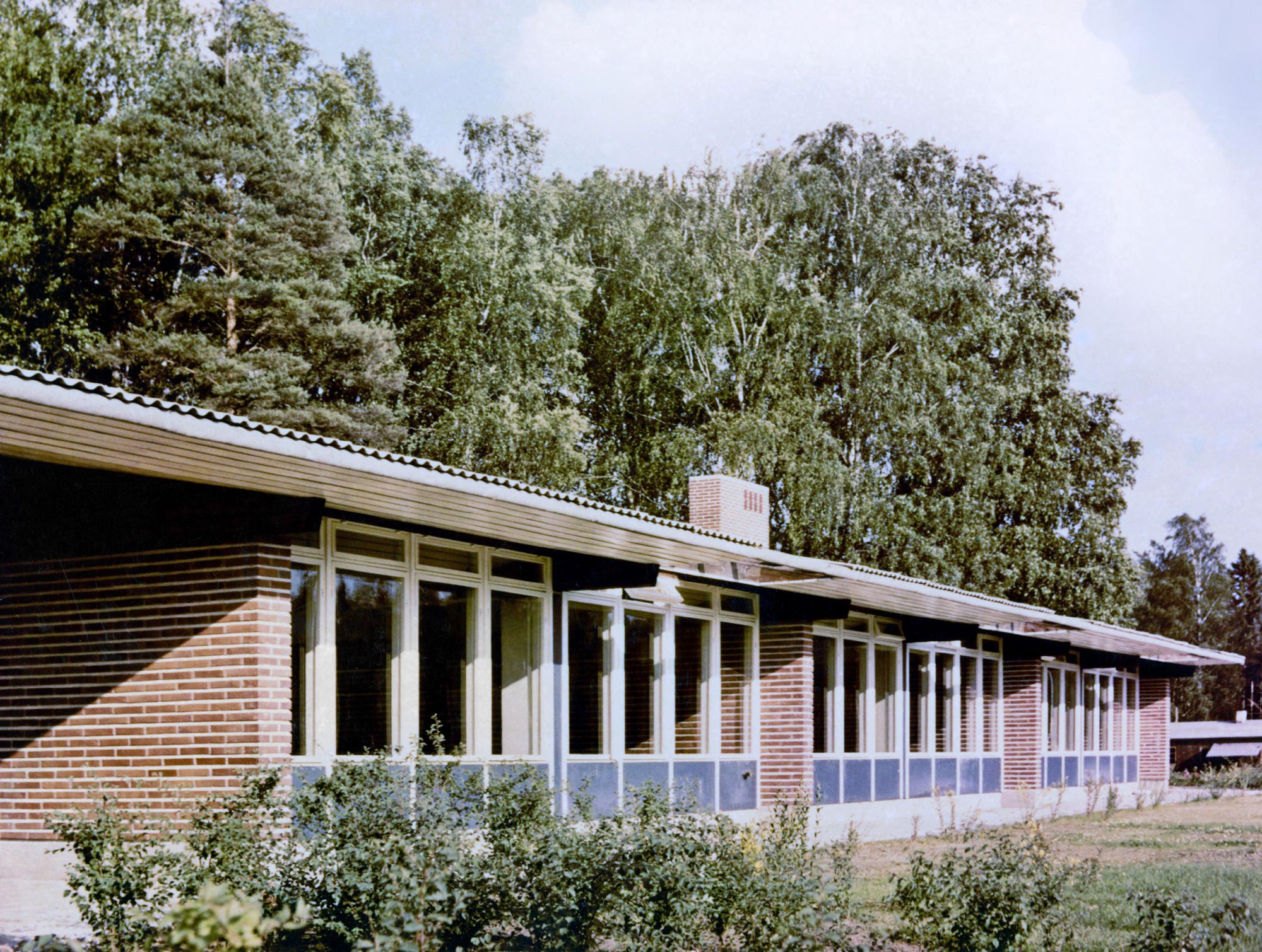 Värivalokuva Tapiolan lastentalosta Espoossa. Rakennus on yksikerroksinen, pitkä ja matala, tiiliverhoiltu seinä ja suuret ikkunat, joiden alareunassa on siniset paneelit. Katto on loiva ja harmaa, ja keskellä näkyy tiilinen savupiippu. Taustalla on tiheä vihreä puusto ja etualalla pensaita ja nurmikkoa kirkkaassa päivänvalossa.