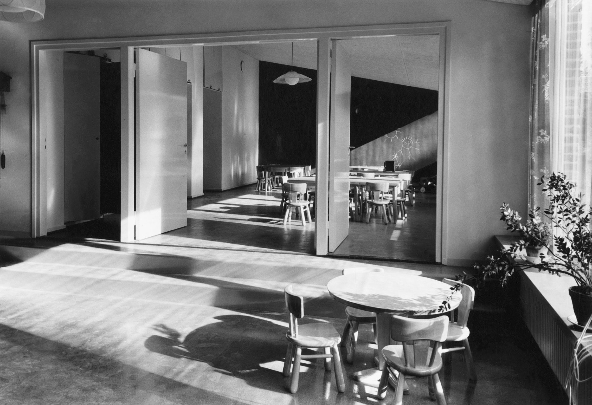 Mustavalkoinen valokuva Maunulan lastentarhan sisätilasta. Etualalla on pieni pyöreä pöytä ja neljä lasten tuolia, joihin osuu kirkas auringonvalo suurista ikkunoista oikealla. Taustalla näkyy avattujen pariovien takana toinen tila, jossa on useita pieniä pöytiä ja tuoleja ryhmissä. Seinällä on tumma varjo ja katossa riippuu valaisimia. Oikeassa reunassa ikkunalaudalla on viherkasveja.