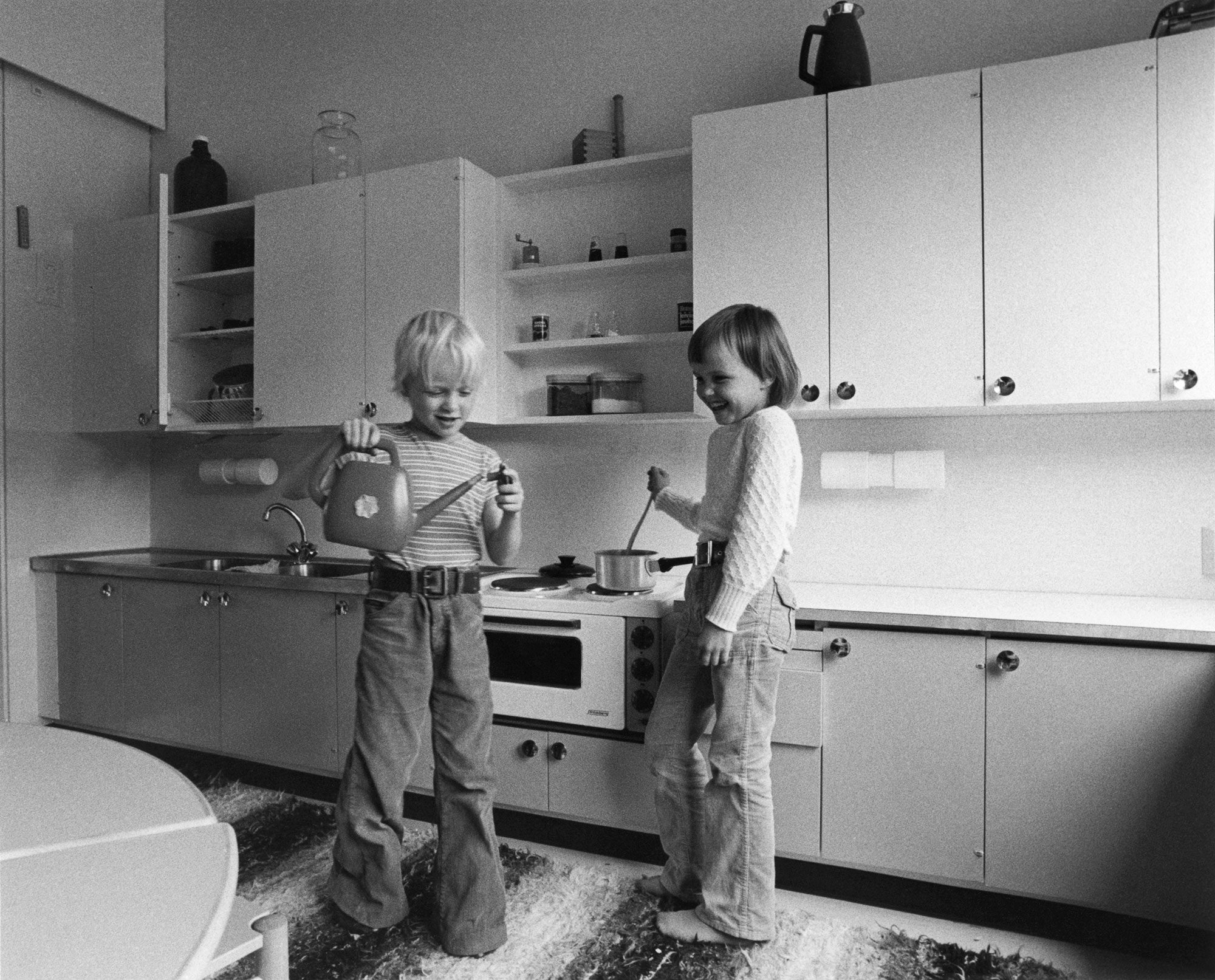 Mustavalkoisessa valokuvassa kaksi lasta seisoo keittiönurkkauksessa, jossa on vaaleat kaapistot, avohyllyjä ja tiskiallas vasemmalla. Yksi lapsi pitää kädessään kastelukannua, toinen sekoittaa kattilaa lieden päällä. Etualalla näkyy pöydän reuna, ja lattialla on kuvioitu matto.