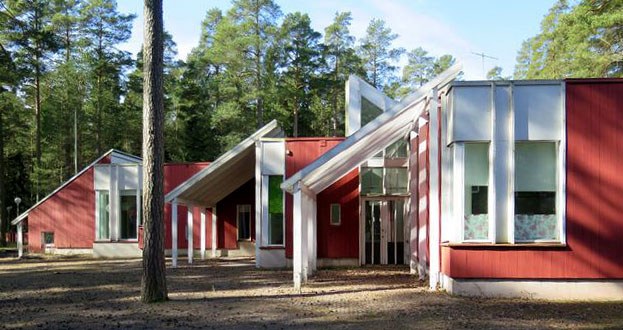 Värivalokuva Länsi-Säkylän päiväkodista metsäisessä ympäristössä. Rakennus on punainen, puurakenteinen ja siinä on useita viistokattoisia osia, jotka muodostavat rytmikkään kokonaisuuden. Valkoiset katosrakenteet ja suuret ikkunat korostavat sisäänkäyntejä. Taustalla näkyy tiheä mäntymetsä ja etualalla hiekkainen piha, jossa kasvaa yksittäisiä puita.