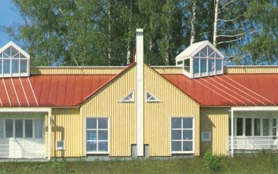 Vaaleankeltainen, yksikerroksinen rakennus, jossa on punainen harjakatto ja kaksi suurta kattoikkunaa. Rakennuksen keskellä on valkoinen savupiippu ja kolmionmuotoisia ikkunoita. Etuosassa on useita suorakaiteen muotoisia ikkunoita ja ovia, ja taustalla näkyy vihreitä puita kirkkaassa päivänvalossa.