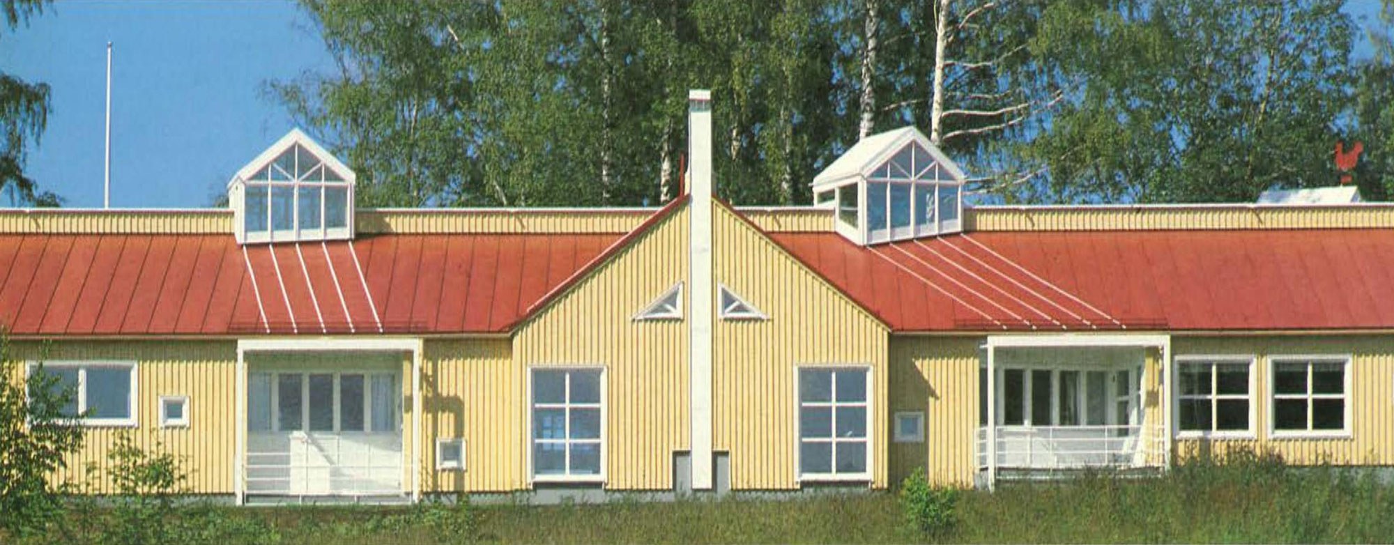 Vaaleankeltainen, yksikerroksinen rakennus, jossa on punainen harjakatto ja kaksi suurta kattoikkunaa. Rakennuksen keskellä on valkoinen savupiippu ja kolmionmuotoisia ikkunoita. Etuosassa on useita suorakaiteen muotoisia ikkunoita ja ovia, ja taustalla näkyy vihreitä puita kirkkaassa päivänvalossa.