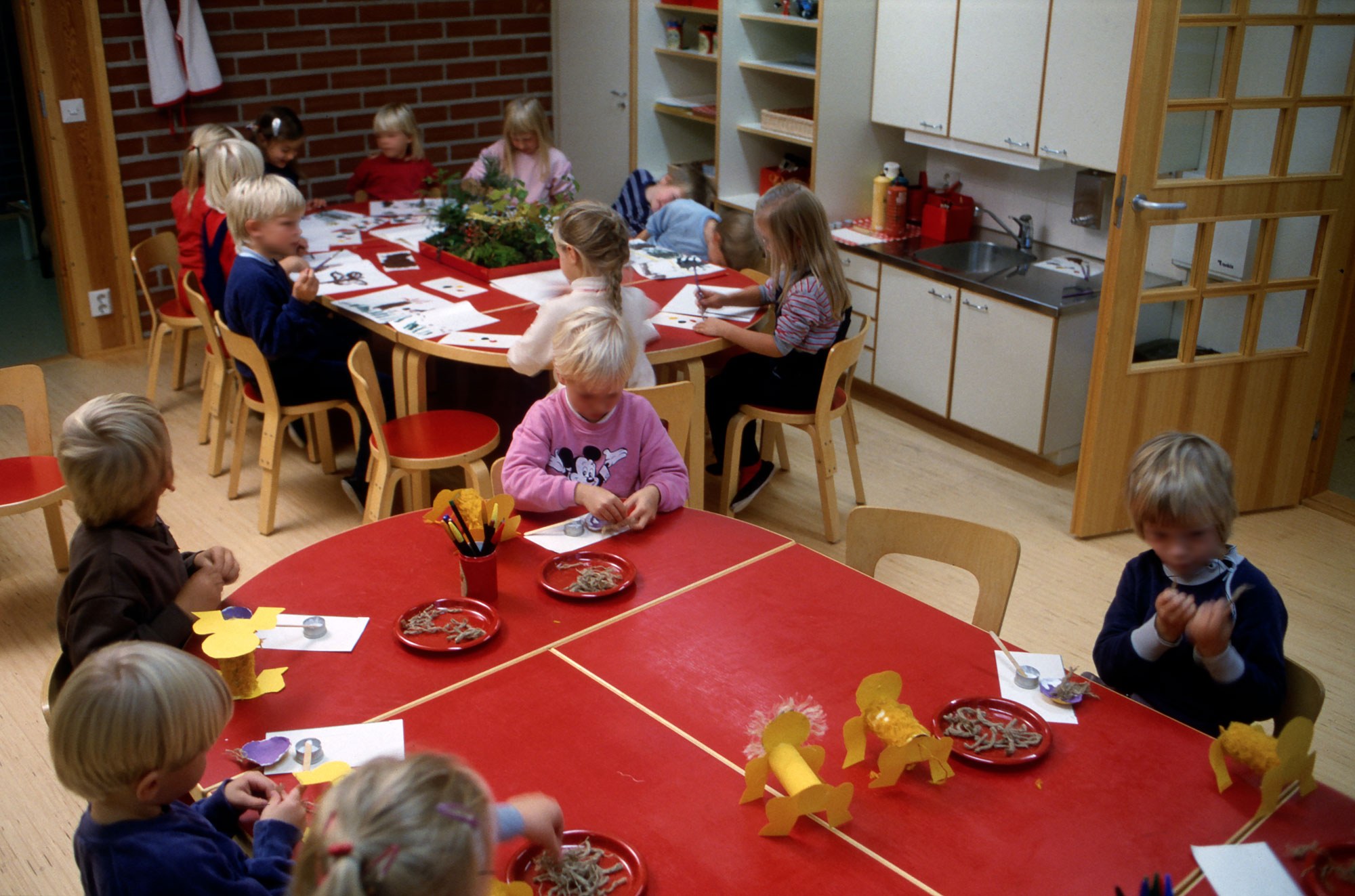 Värivalokuva päiväkodin sisätilasta, jossa useita lapsia istuu punaisten pöytien ääressä askartelemassa. Pöydillä on värikkäitä paperikoristeita, punaisia lautasia, kyniä ja askartelumateriaaleja. Taustalla näkyy toinen pöytäryhmä, kaapistoja, avohyllyjä ja pieni keittiönurkkaus, jossa on punaisia astioita. Oikealla on lasiovi ja ikkunasta tulee luonnonvaloa.