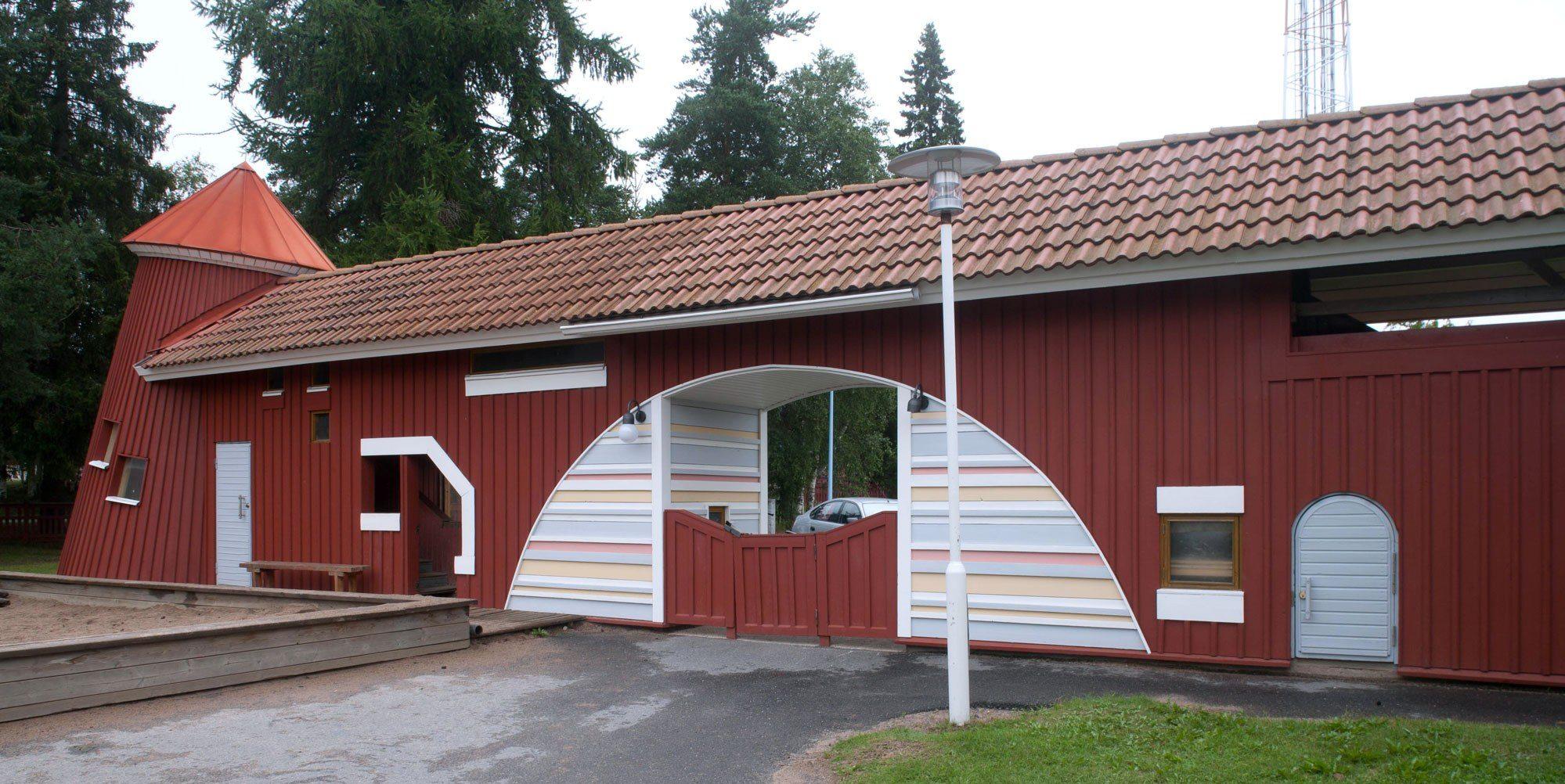 Värivalokuva Sailolan päiväkodista Oulussa. Rakennus on punainen, puurakenteinen ja siinä on tiilikatto. Keskellä on suuri kaareva portti, jonka sisäpinta on koristeltu valkoisilla ja pastellisävyisillä vaakaraidoilla. Vasemmassa päädyssä on torni, jossa on kartiomainen katto, ja seinässä pieniä suorakaiteen ja kaarevia ikkunoita sekä ovia. Etualalla on asfalttipäällysteinen piha, hiekkalaatikko ja nurmikko, taustalla vihreitä puita.